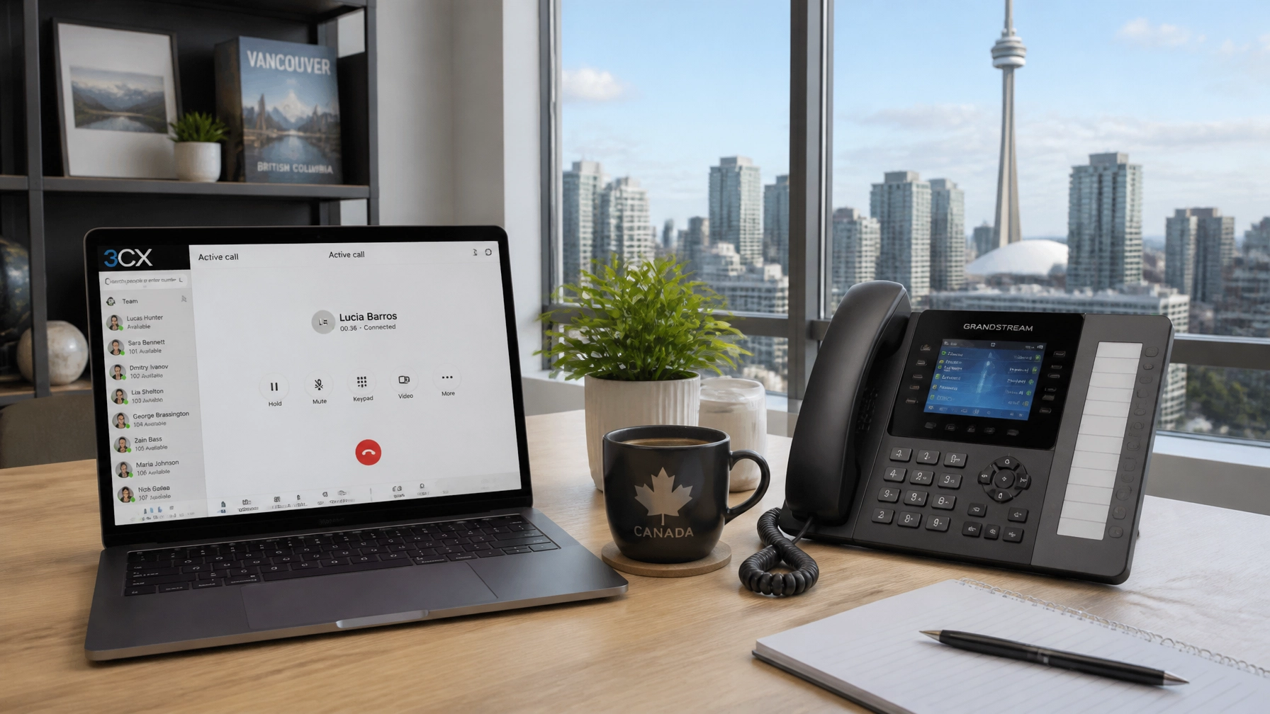 Laptop on a wooden desk showing a video call, with a potted plant, a Canada mug, and an office phone nearby.